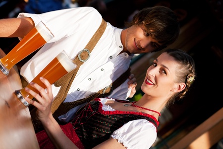 Couple in traditional bavarian tracht dirndl and lederhosen in front of a beer tent at the oktoberfest or in a beer garden enjoying a glass of tasty wheat beer