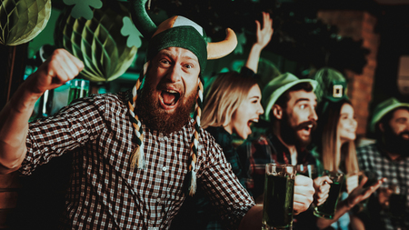 Man in funny hat celebrates st patrick s day bar counter alcohol handling ginger beard smiling male good festive mood bright lights funny club visitor open mouth glass of beer