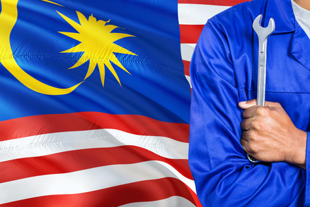 Malaysian mechanic in blue uniform is holding wrench against waving malaysia flag background crossed arms technician