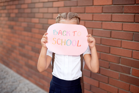 Little girl blonde in school uniform stands near a brick wall holding a blackboard with the text back to school Stock Photo