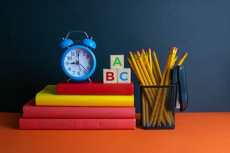 A stack of books a green flower blue clock and a glass of pencils and wood cubes with abc stand on an orange background Stock Photo