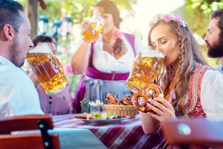 Woman in tracht looking into camera while drinking a mass of beer surrounded by her friends