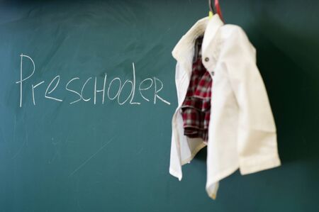 A hanger with school clothes of a small child hangs above the blackboard and the inscription preschooler is on it Stock Photo