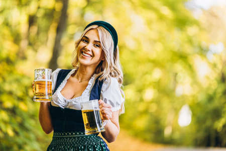 Pretty happy blonde in dirndl traditional festival dress holding two mugs of beer outdoors in the forest with blurred background oktoberfest st patrickâ€™s day international beer day concept