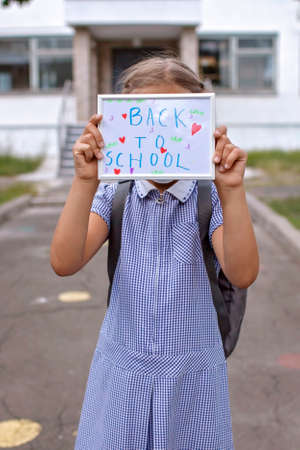 Elementary school girl in medical mask holds picture with back to school message first offline day with social distance rules new normal education reunion after lockdown and quarantine Stock Photo