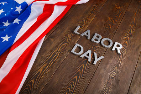 Words labor day laid with silver metal letters on wooden surface with crumpled usa flag on left side in diagonal perspective view