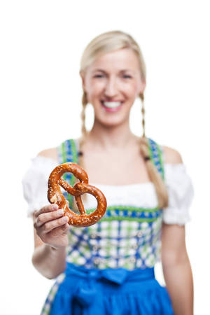 Beautiful smiling young blond woman in a dirndl holding a pretzel or salted glazed brittle german biscuit in the shape of a knot in her hand isolated on white