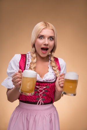 Half length portrait of young surprised blonde wearing pink dirndl and white blouse holding in both hands beer mugs looking at us isolated on dark background Stock Photo