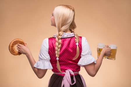 Half length portrait of young blonde wearing pink dirndl and white blouse standing back to us holding tasty bagel in one hand and beer mugs in another isolated on dark background