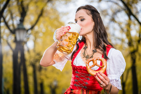 Girl in traditional bavarian tracht drinking beer out of a huge mug holding pretzel in other hand