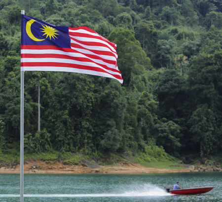 Malaysia flag jalur gemilang waving with the background of boat at the lake and malaysian rainforest trees Stock Photo