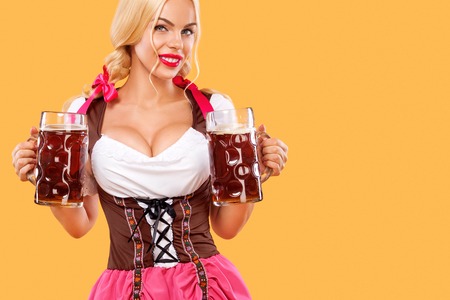 Young oktoberfest girl waitress wearing a traditional bavarian dress serving big beer mugs on orange background