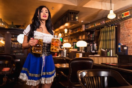 Beautiful female waitress wearing traditional dirndl and holding huge beers in a pub