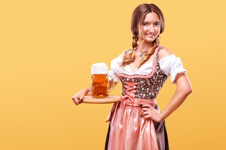 Young oktoberfest girl waitress wearing a traditional bavarian dress serving big beer mugs on orange background with copy space