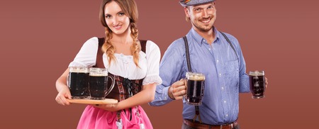 Portrait of happy oktoberfest man and woman wearing a traditional bavarian clothes serving big beer mugs on brown background Stock Photo