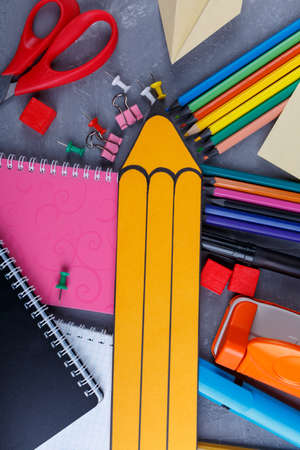 A large yellow paper pencil next to a variety of multi colored pencils several notebooks clamps and crayons and other office supplies view from above